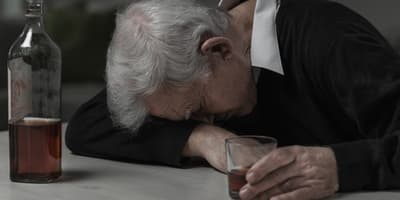Elderly man asleep at table holding glass of alcohol